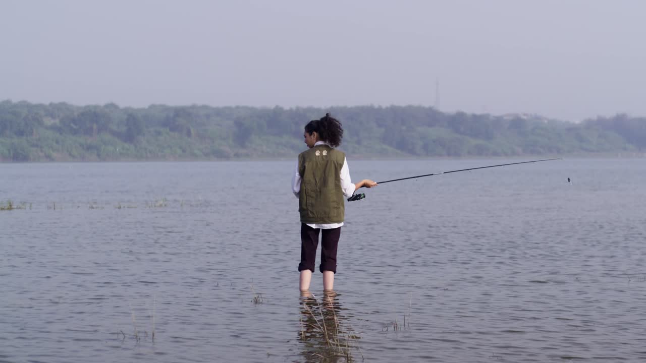 Seorang gadis Asia Selatan mengenakan rompi luar ruangan sedang memancing di air danau yang dangkal, terlihat dari belakang pada siang hari yang tenang