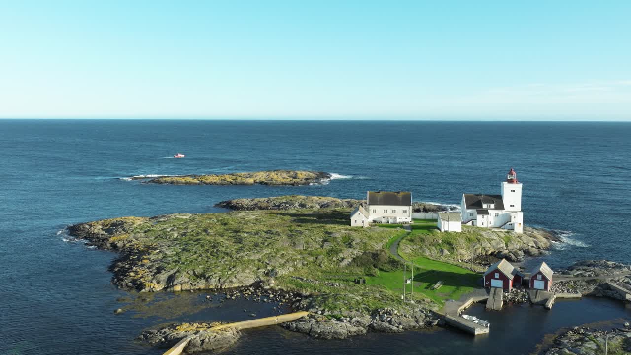 A tranquil scene shows a lighthouse standing tall on a rocky island surrounded by calm blue waters. Nearby buildings add charm to the coastal landscape under a clear sky