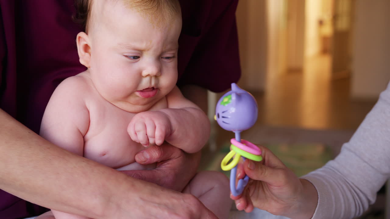 Caregiver holding a baby upright during a developmental exercise while another adult engages with a toy nearby
