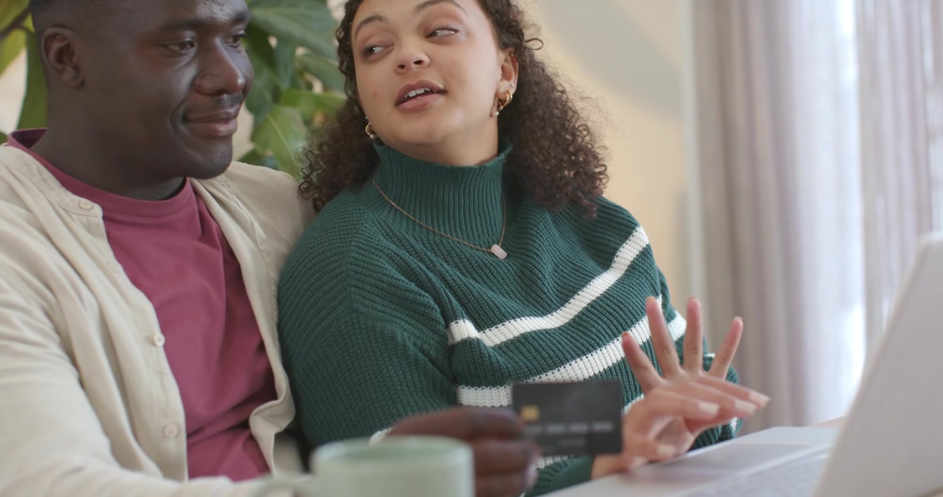 Diverse couple in living room sitting on couch, man offering card, woman typing purchase on laptop