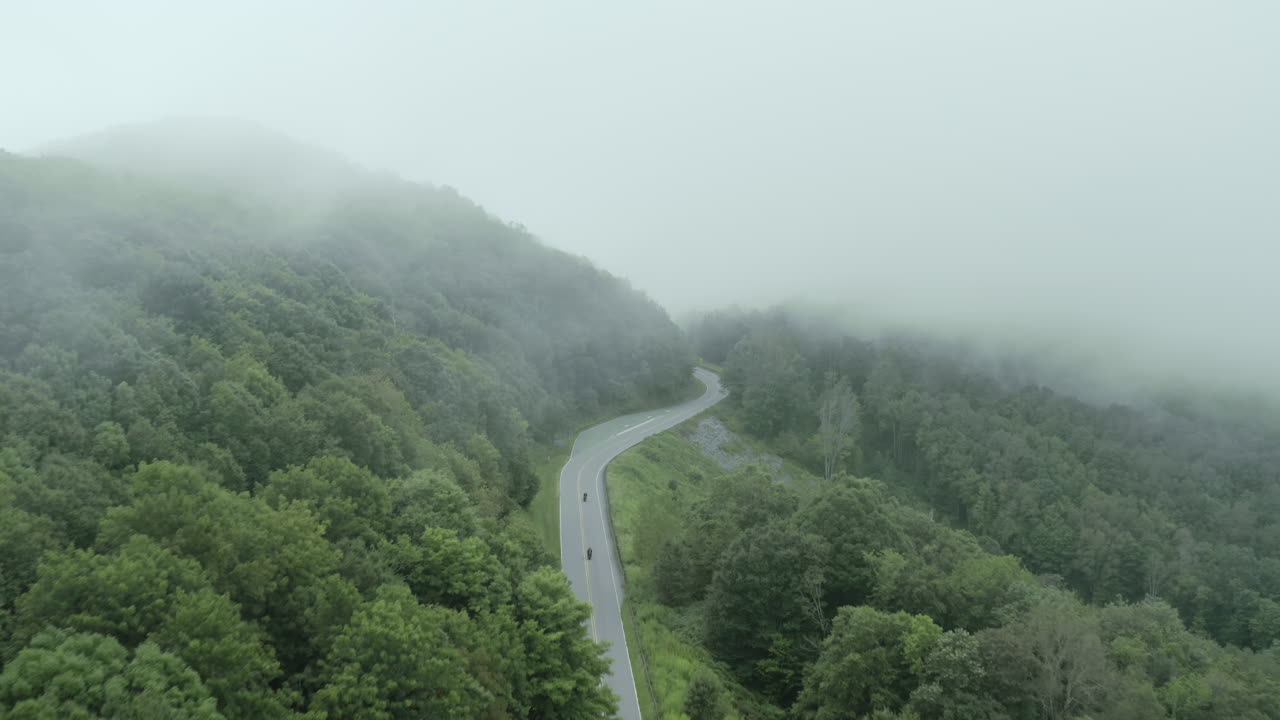 바람이 부는 안개가 자욱한 산악 도로 아래로 빠르게 이동하는 오토바이를 따라가는 공중, 4k
