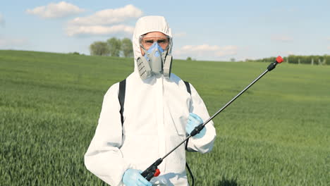 close-up view of caucasian researcher man in white protective suit and goggles holding pulverizator in the green field while looking at camera