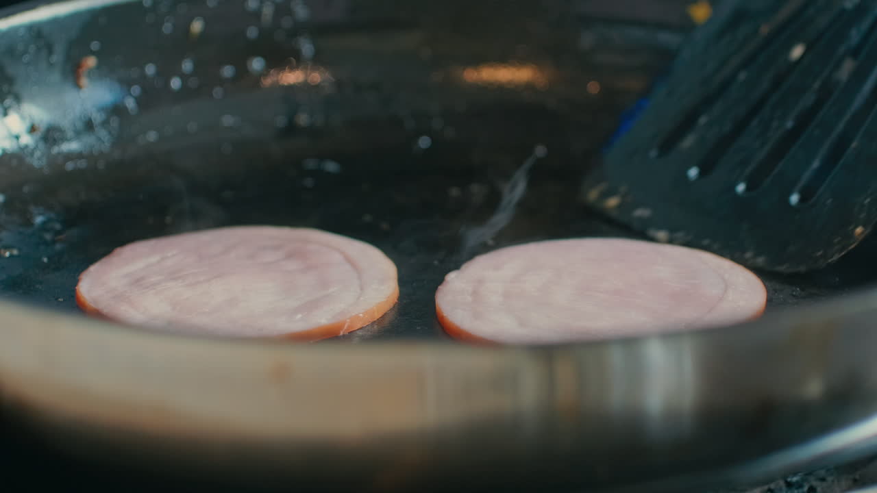 Close-up of a spatula flipping slices of Canadian bacon or ham frying in a hot skillet during breakfast preparation