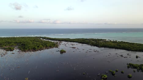 A-drone-slowly-flies-bacwards-above-a-lagoon-and-tropical-forest-looking-out-towards-the-ocean-and-a-coral-reef-on-the-Cayman-Islands-in-the-Caribbean-at-sunset