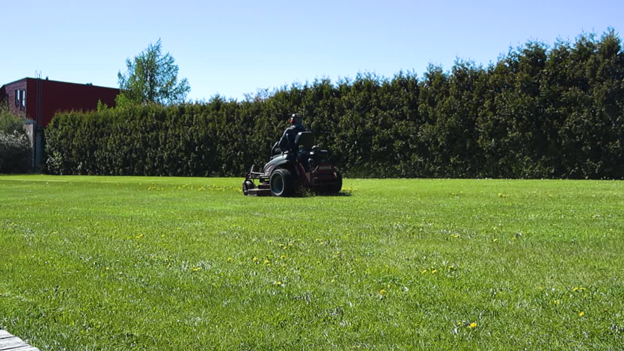 Vibrant and gorgeous footage of a man with safety gear mowing lawn with his high end lawn mower during a sunny day at a countryside garden. Grass is flying out from the machine in slow motion.