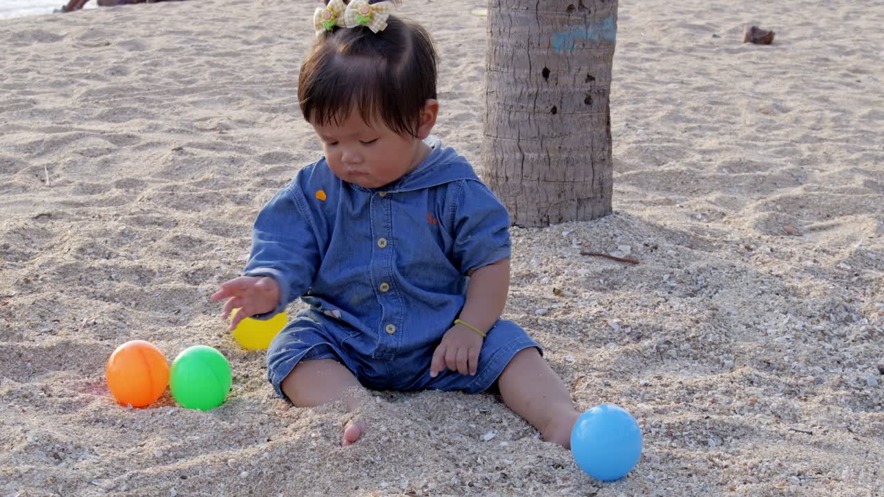 A joyful toddler enjoys a playful moment the beach surrounded by colorful balls in the sand.