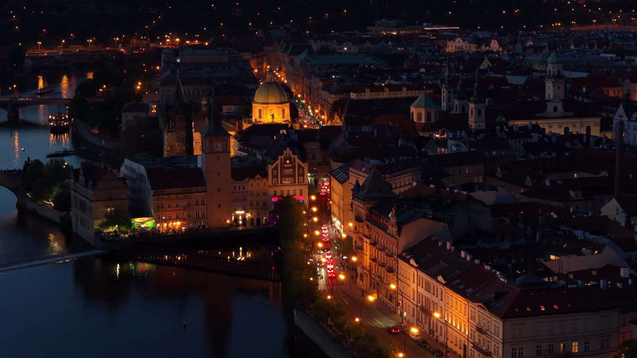 Vista aerea con drone della Città Vecchia di notte, con strade illuminate, una cupola dorata e guglie di chiese lungo la riva del fiume Moldava.