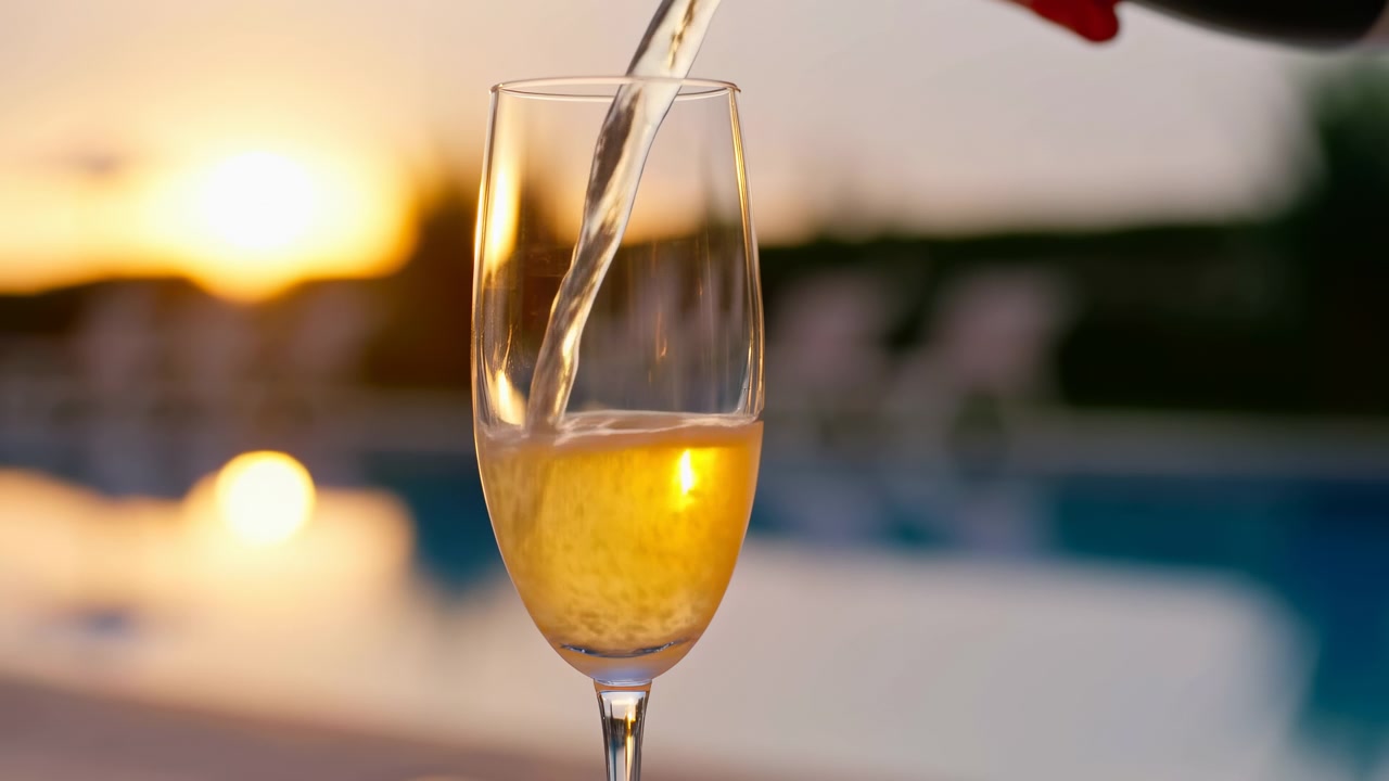 Primo piano di vino spumante che scorre in un alto bicchiere a flûte con una calda luce del tramonto e uno sfondo sfocato di una piscina esterna.