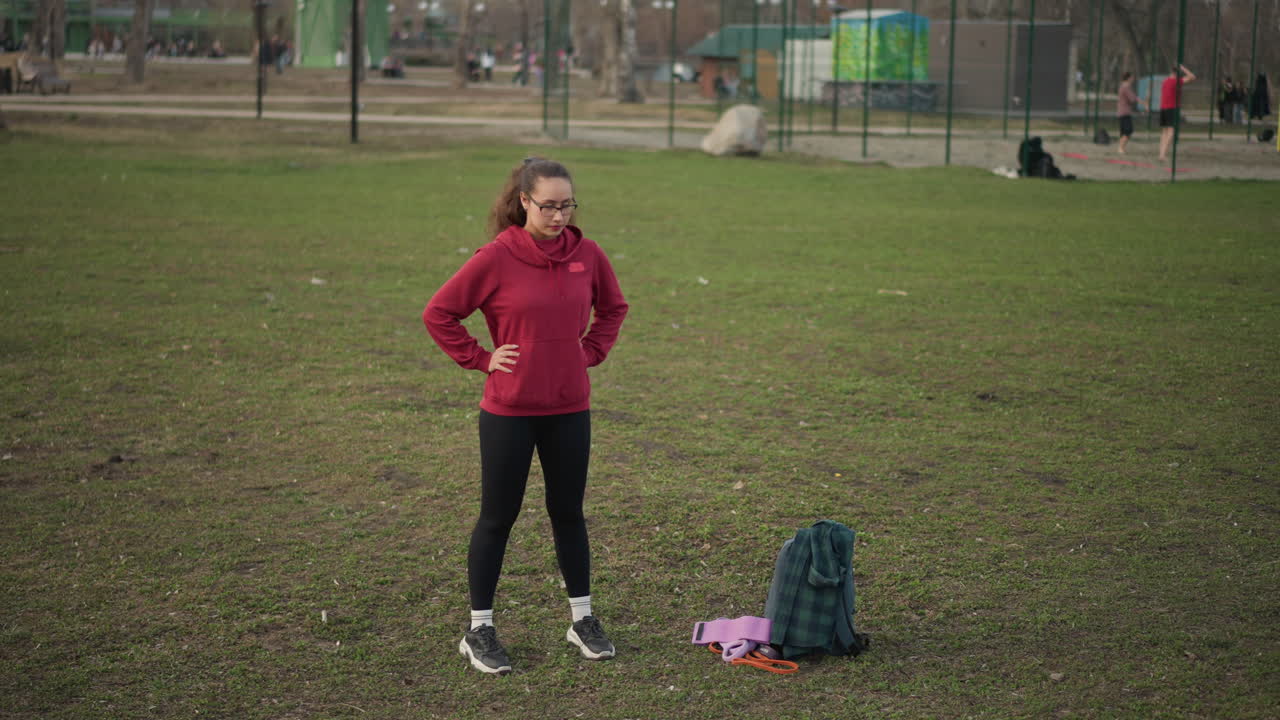 Female Exercising In Urban Park Environment, Woman Performing Warmup Stretch In Park Under Cloudy Sky, Person Dressed In Hoodie Engaging In Mobility Exercises On City Park Grass Field