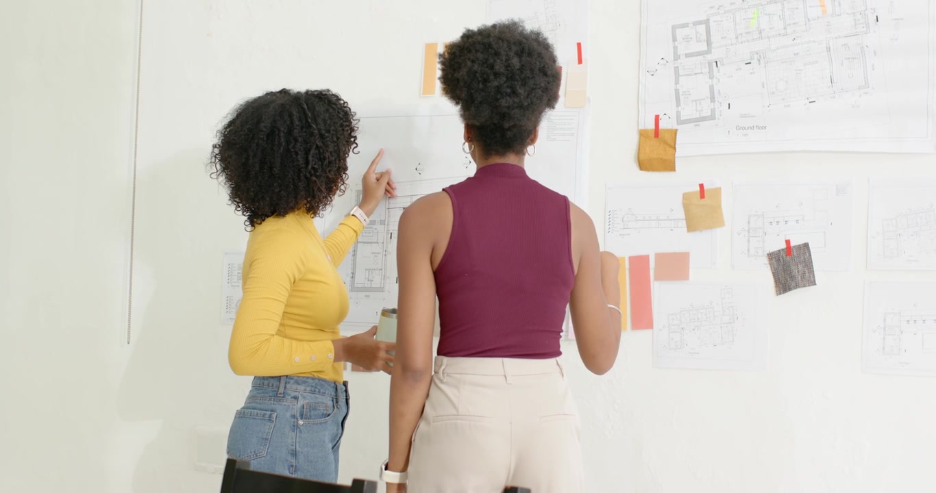 Diverse female designers reviewing plans at pin-up wall, pointing with pencil, moving swatches