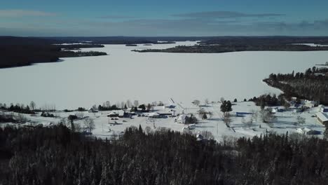 Frío-Y-Soleado-Cielo-Azul-Día-De-Invierno-En-La-Campiña-Del-Norte-De-Canadá---Drone-4k-Antena-Pequeña-Ciudad-Junto-A-Un-Lago-Congelado