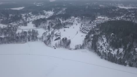 Vista-De-Gran-Angular-Con-Cámara-Lenta-Mirando-Hacia-Abajo-Sobre-El-Paisaje-Invernal-Con-Una-Pequeña-Comunidad-Situada-Muy-Cerca-De-Un-Gran-Lago