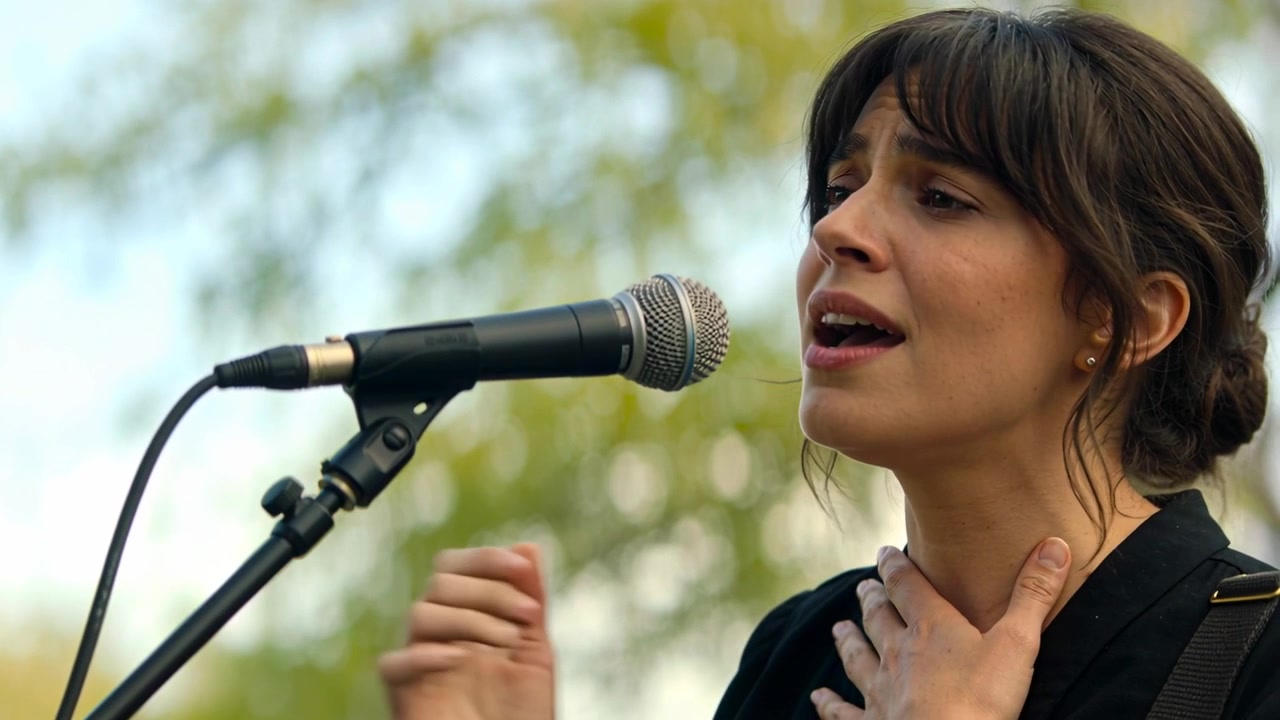 Cantante che si esibisce su un palco all'aperto, toccandosi la gola e gesticolando vicino al microfono, fogliame.