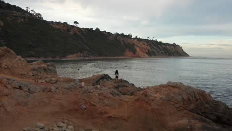 Eine-Frau-Steht-Am-Strand-In-Der-Nähe-Von-Rancho-Palos-Verdes-Und-Beobachtet-Die-Surfer-Bei-Sonnenuntergang---Luftparallaxenumlaufbahn