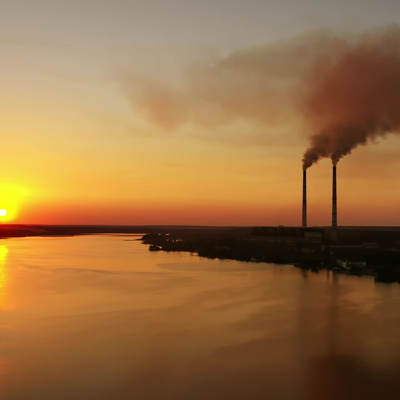 Industrial plant near the evening river. Dirty smoke from pipes goes in the air against the setting sun. Silhouette of a factory near the lake on orange sky background.
