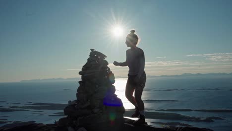 norwegian woman building mountain of stones by the coast with bright sunlit background - wide shot