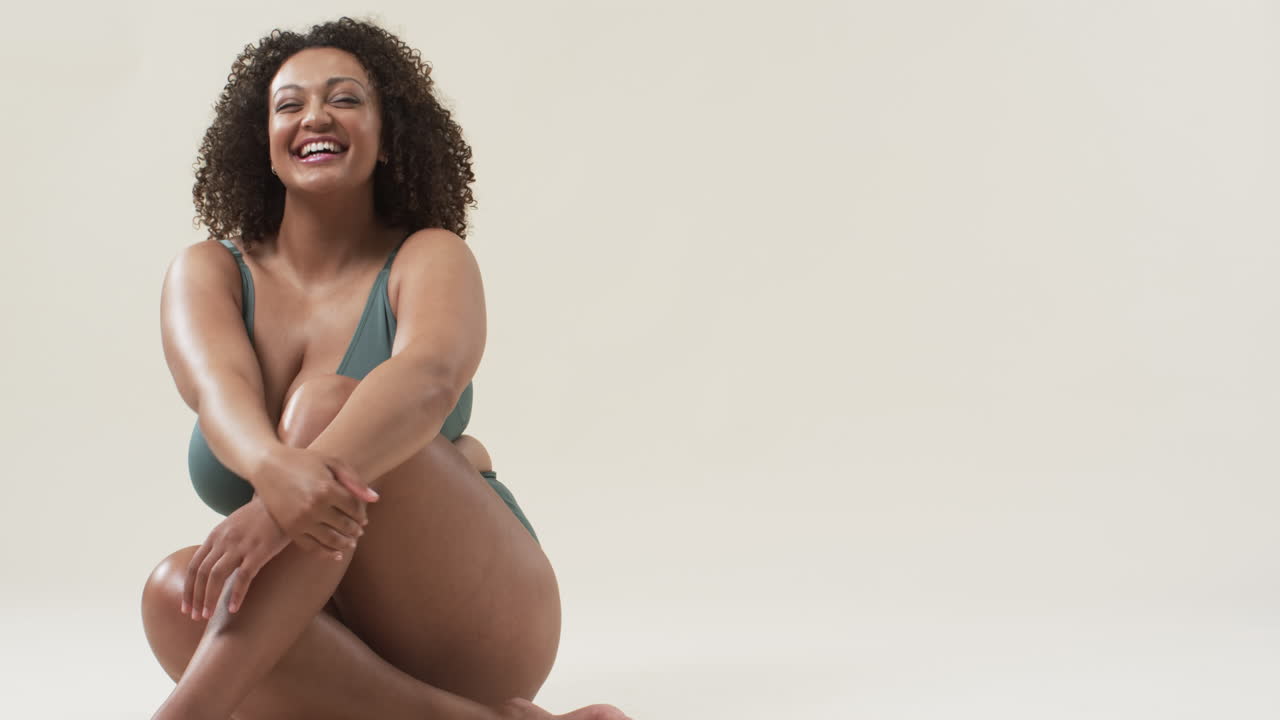 A plus size woman with curly hair wearing green bikini is sitting on floor, smiling, copy space