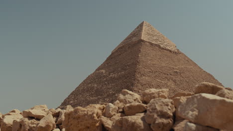wide shot of the great pyramid of giza in egypt on a sunny clear day. the pyramid stands tall with sandy rocks in the foreground, blue sky above, and desert scenery around this famous world landmark