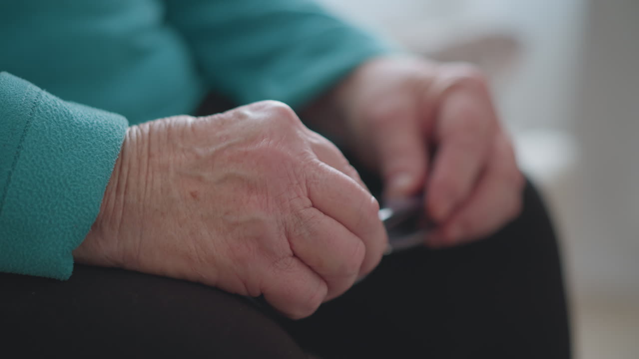 Elderly woman in green teal sweater sitting down, adjusting glasses with hands, close-up focus on hands and glasses, soft lighting with light background, cozy indoor setting