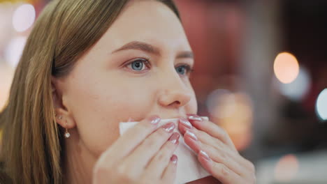 close-up of lady cleaning mouth with napkin while sitting in mall dining area, soft blur background with tables, chairs, and people passing by