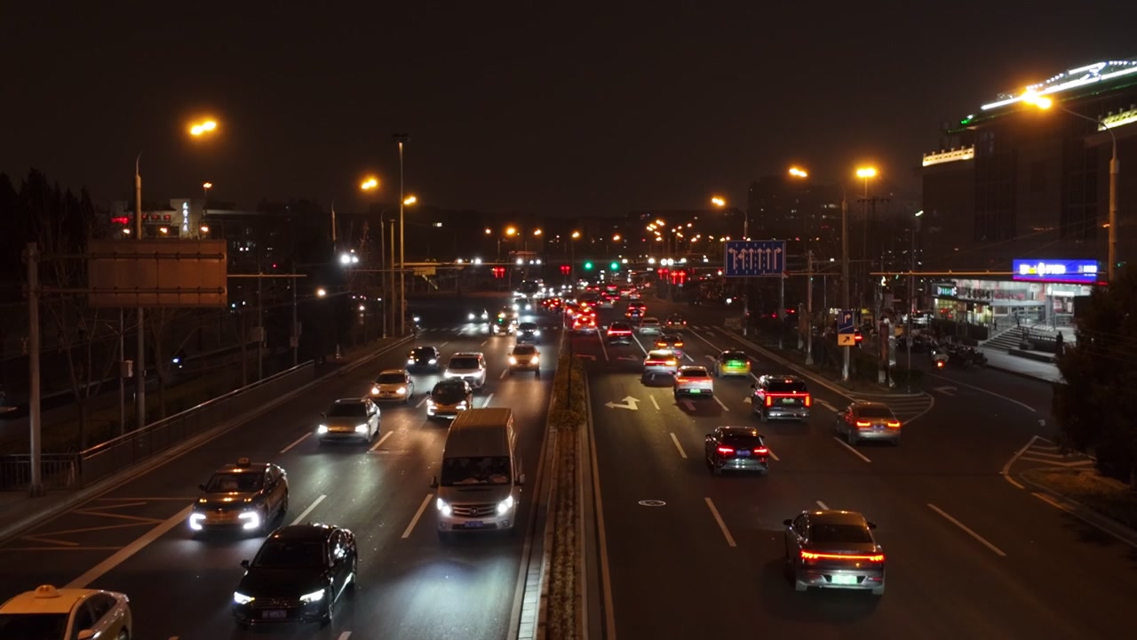 Motorvejstrafik strømmer gennem Beijing om natten med oplyste køretøjers baglygter og forlygter. Et hævet statisk skud fanger flere baner af biler og busser under gadelamper i et bymiljø.