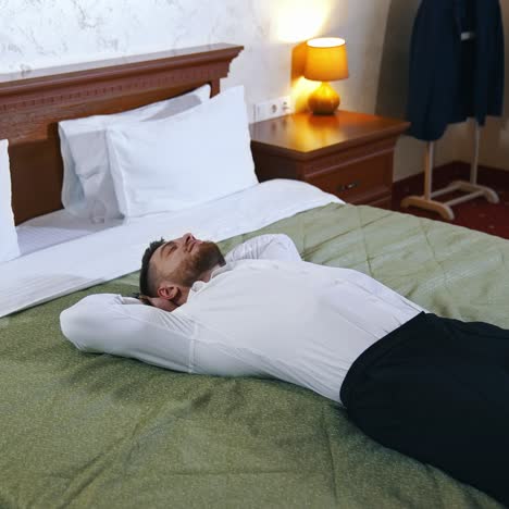 Businessman rest at daytime. Handsome man in white shirt and black trousers lying on bed in comfortable hotel. Adult male relaxing during business trip