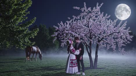 Romantic couple wearing traditional clothes embracing under a blooming cherry blossom tree in a foggy meadow at night, with a full moon and a horse in the background