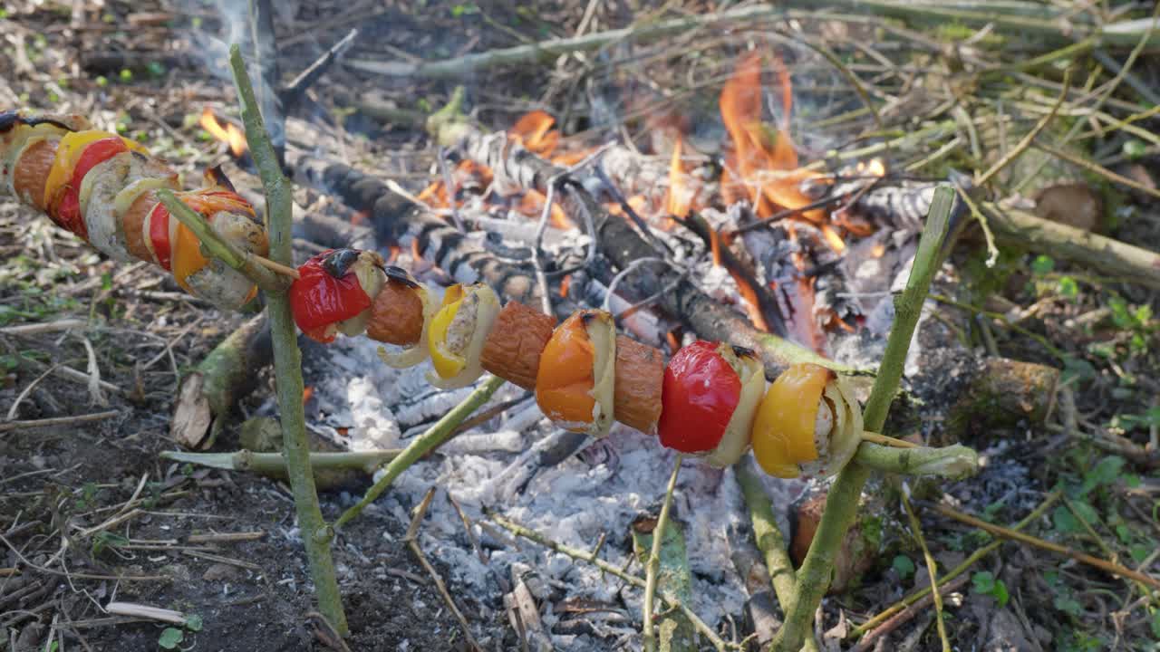 A skewer with vegetables and sausages roasting over a campfire in nature