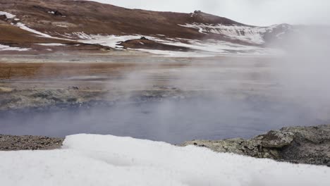 Tiro-Bajo-Sobre-La-Nieve-De-Una-Piscina-De-Barro-Azul-Oscuro-Que-Forma-Vapor-Blanco-En-Islandia