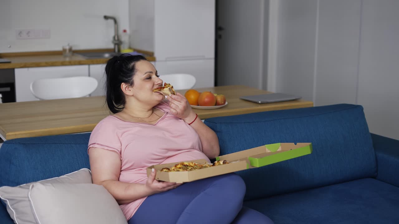 Donna curvy che mangia una fetta di pizza con grande piacere, seduta su un divano.