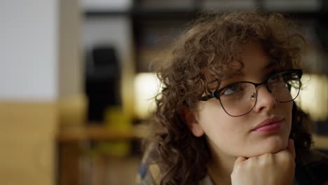 primer plano de una estudiante pensativa con gafas y cabello rizado mira hacia un lado mientras piensa en sus clases en la universidad