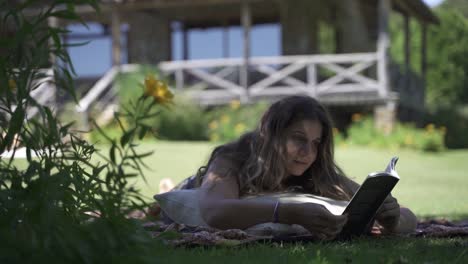 Joven-Mujer-Latina-Sonriendo-Leyendo-Un-Libro-Al-Aire-Libre-En-El-Césped-Con-Vegetación-Y-Fondo-De-Casa-A-Cámara-Lenta-60fps