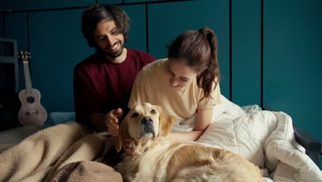 Happy-couple-together-with-their-pet:-A-guy-and-a-girl-play-with-their-dog-of-light-coloring-in-the-bed-of-their-house-against-the-background-of-a-turquoise-wall