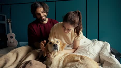 happy couple together with their pet: a guy and a girl play with their dog of light coloring in the bed of their house against the background of a turquoise wall