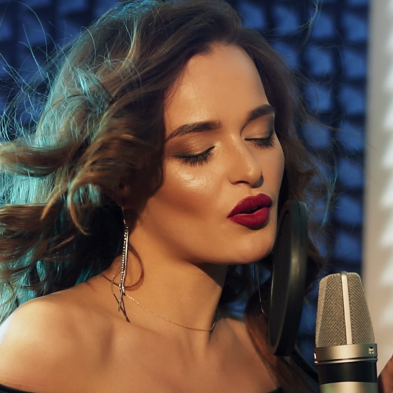 Attractive female with bare shoulders is singing in the professional studio. Beautiful model sings passionately with closed eyes to the modern microphone. Slow motion.