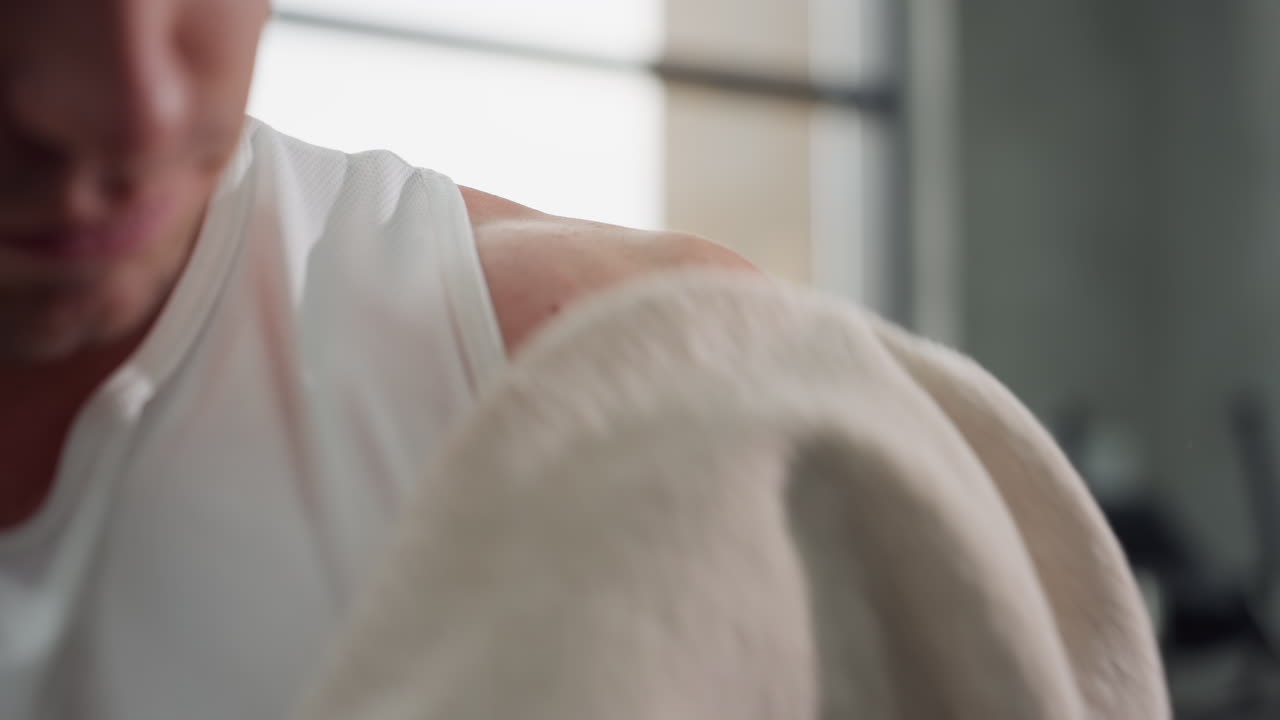 Gym goer presses towel against shoulder to wipe sweat and clean skin after intense workout capturing close up motion and muscle detail under bright natural light in modern fitness environment