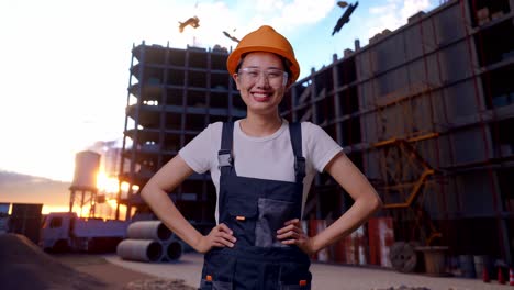 trabajadora asiática con gafas y casco de seguridad de pie con los brazos akimbo mirando a la cámara y sonriendo en el sitio de construcción