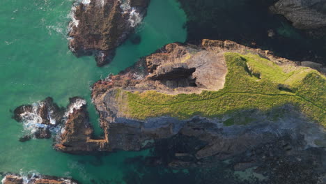 Sea-cliffs-along-Cornwall-coastline-jutting-into-clear-ocean,-aerial-overhead