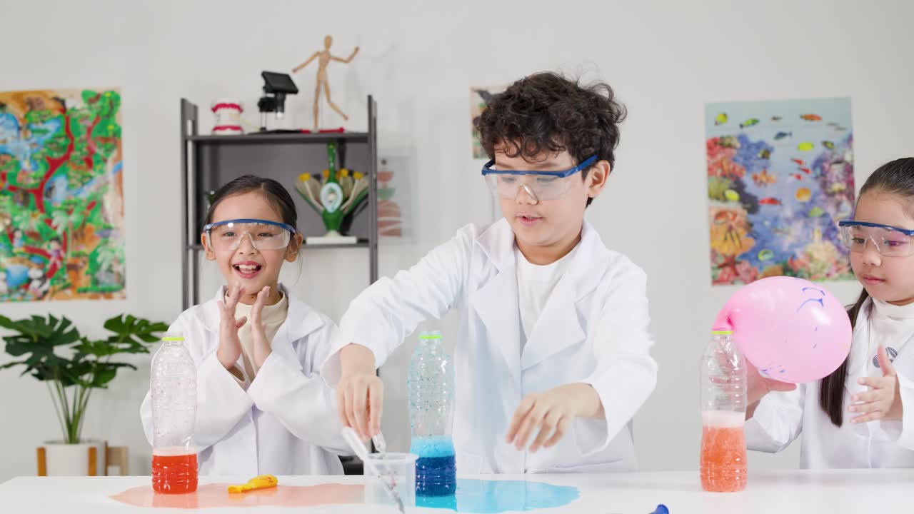 白衣を着たアジア系の学生3人が、カラフルな風船を膨らませる化学反応実験を行う