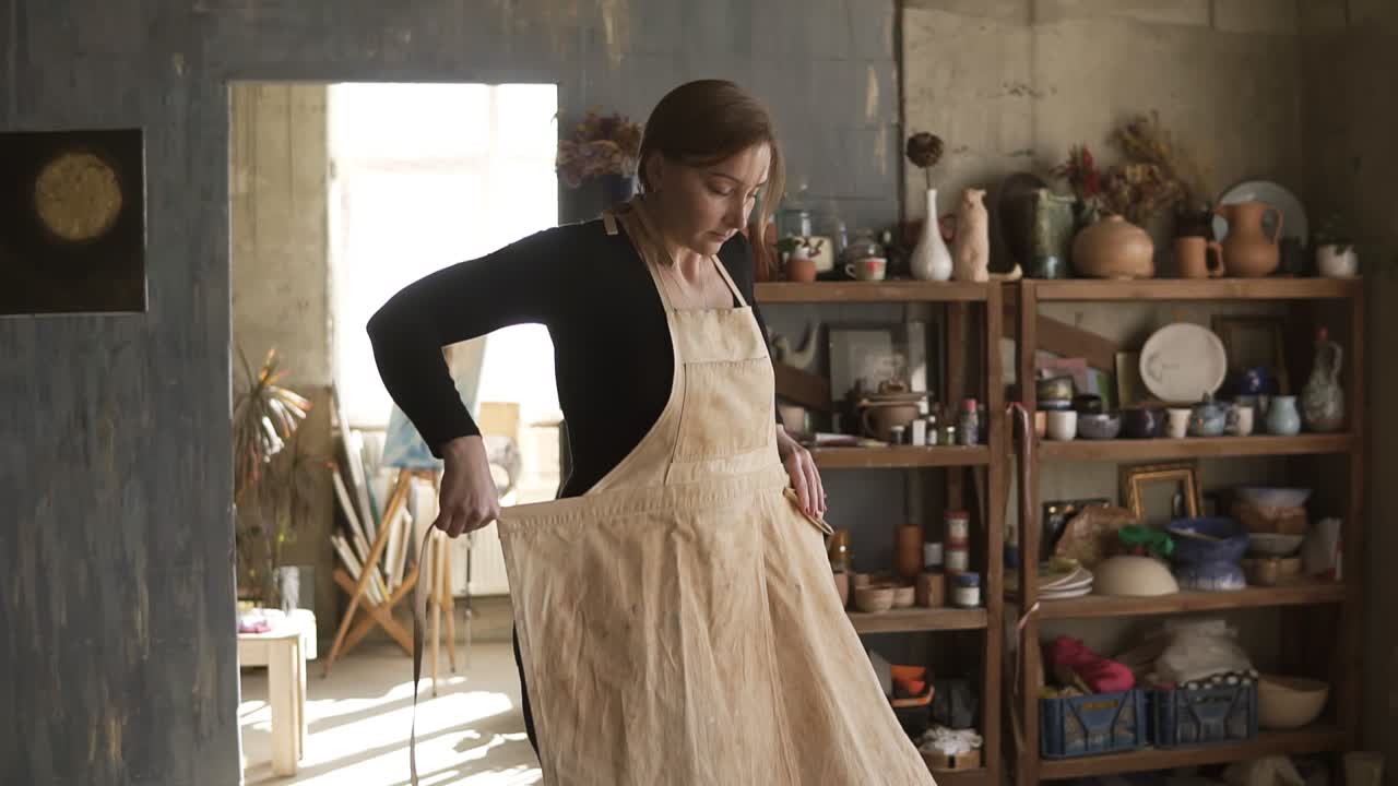 Slow motion footage of young caucasian woman in casual clothes standing in pottery workshop, with ceramic wares and supplies on shelves and table with tools in middle, and putting on beige apron