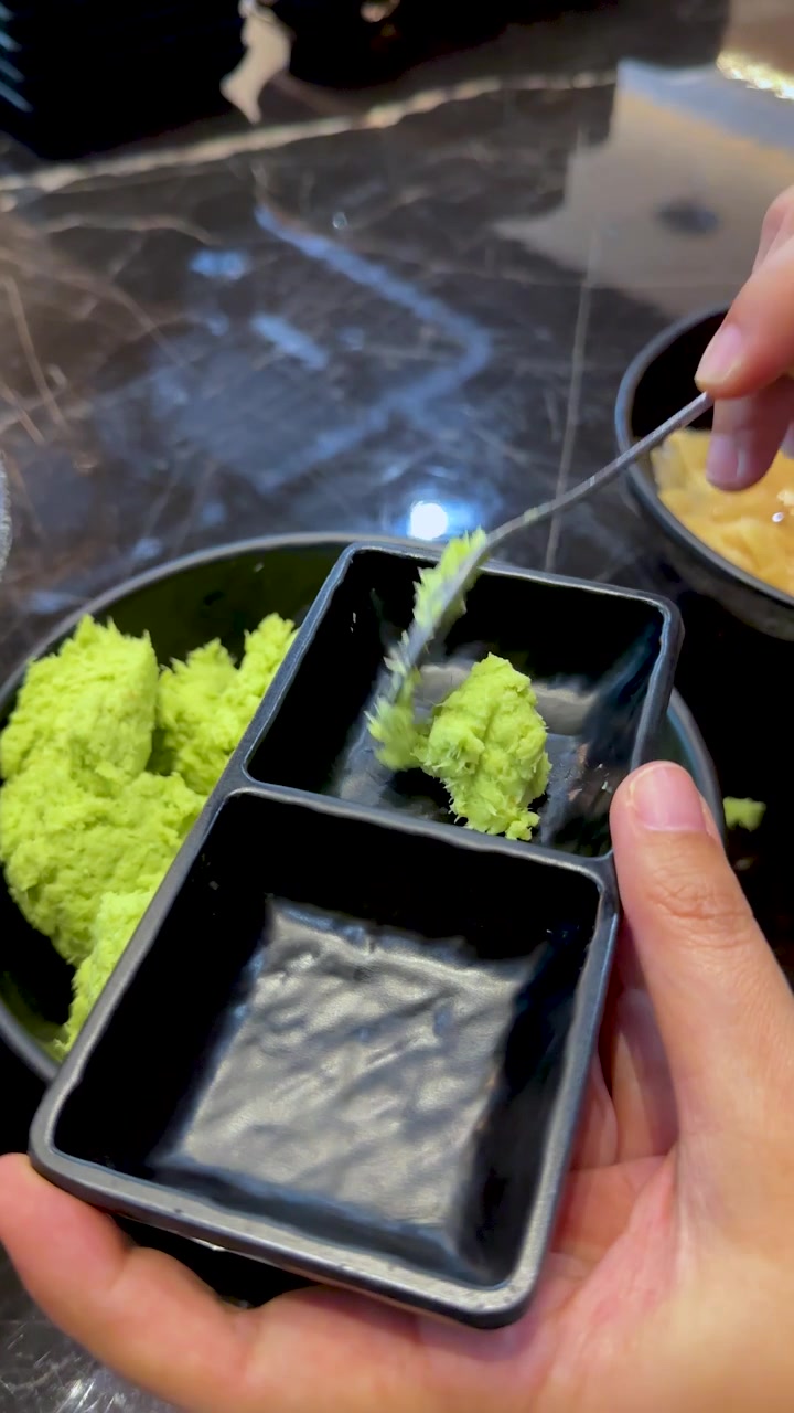 Hand scoops wasabi paste into small black sauce dish at self-serve buffet station
