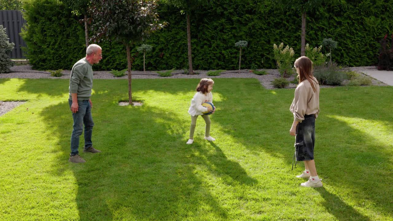 Active Grandfather Throws Ball While Playing With Two Grandchildren In Backyard