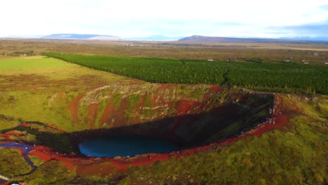drone-shot-of-a-crater-in-Iceland-with-red-earth,-a-small-lake-of-water-inside,-a-little-greenery-above,-a-forest-in-the-background,-and-a-large-plain-in-the-background,-cloudy-but-sunny-sky