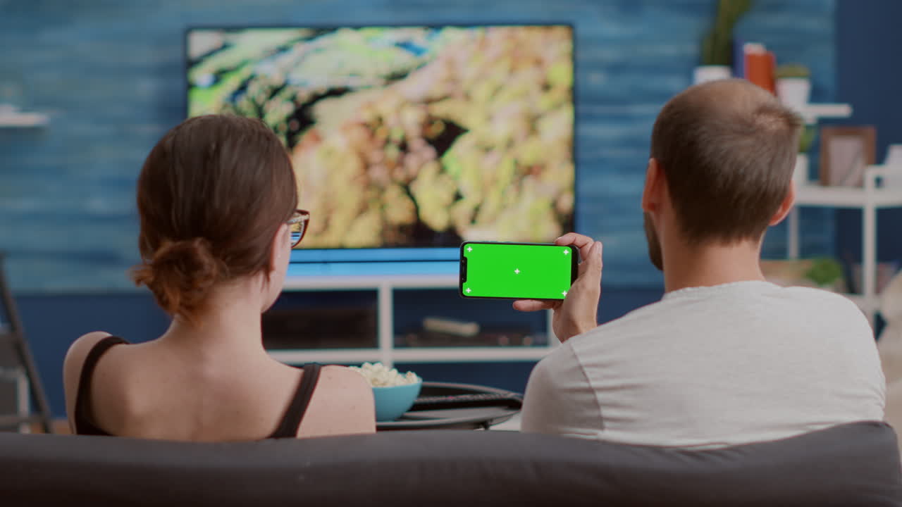 Couple at home watching online tutorials on green screen smartphone while enjoying free time