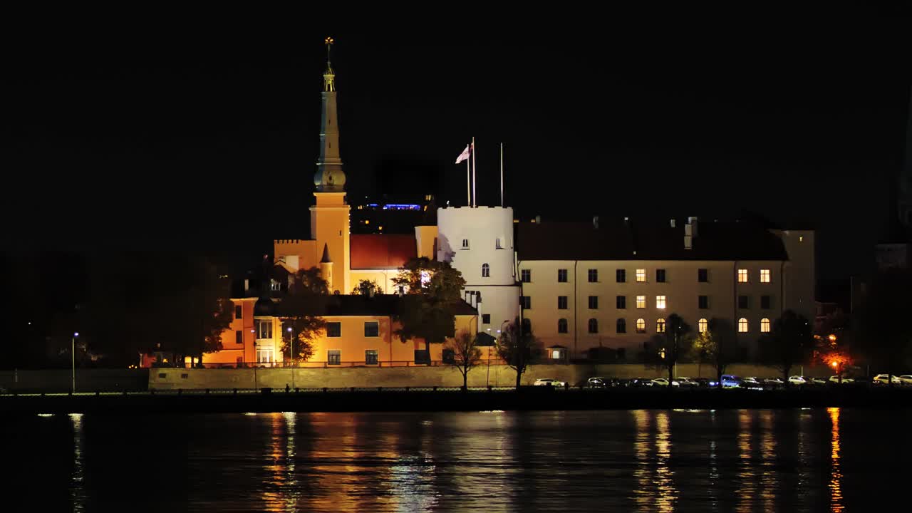 Handheld night scene of Riga Castle glowing beside river with autumn reflections