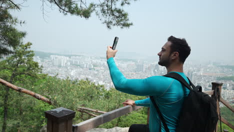 Hombre-Viajero-Tomando-Fotos-En-El-Teléfono-Móvil-En-La-Montaña-Gwanaksan,-Ciudad-De-Seúl-En-Segundo-Plano