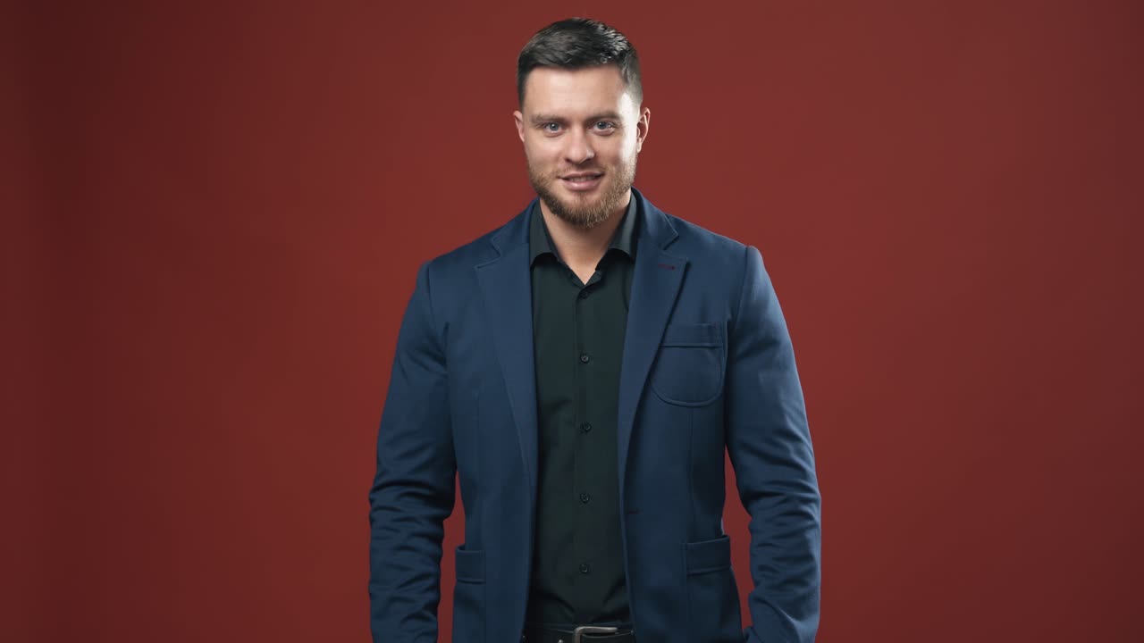 Handsome man in stylish suit. Portrait of young businessman in the red studio. Smiling bearded man in black shirt and blue jacket looking at camera.
