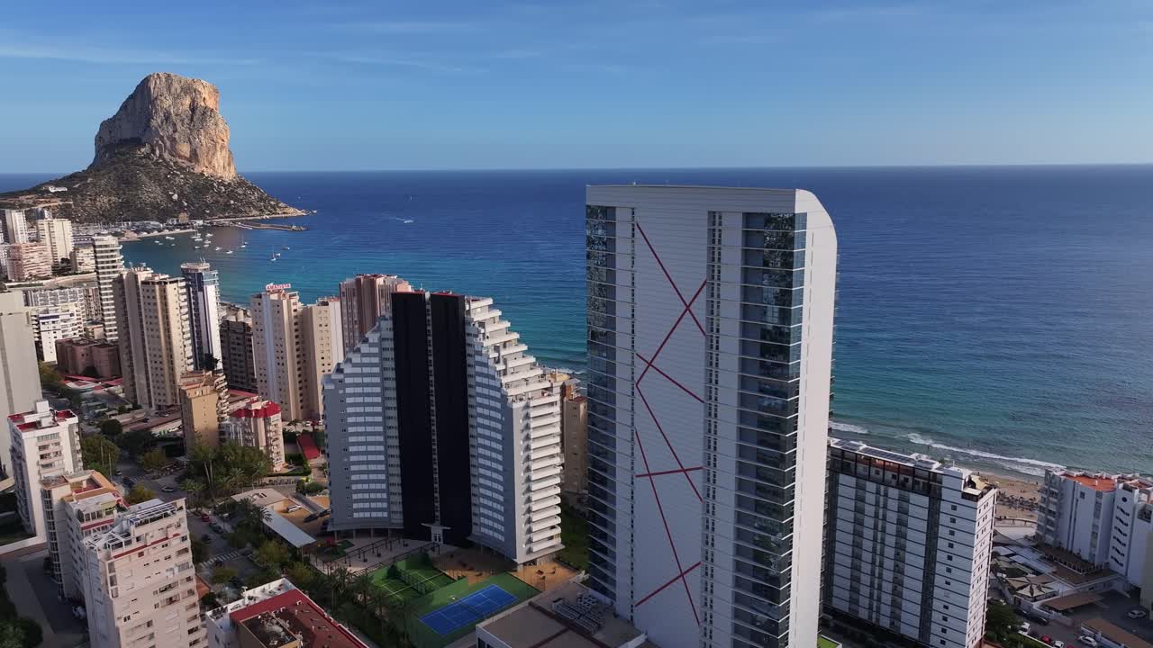 Drone orbiting shot over Calp, Spain shows Peñón de Ifach rising from the sea, with coastal high-rises and the blue Mediterranean stretching into the horizon, warm sunlight and urban grid below