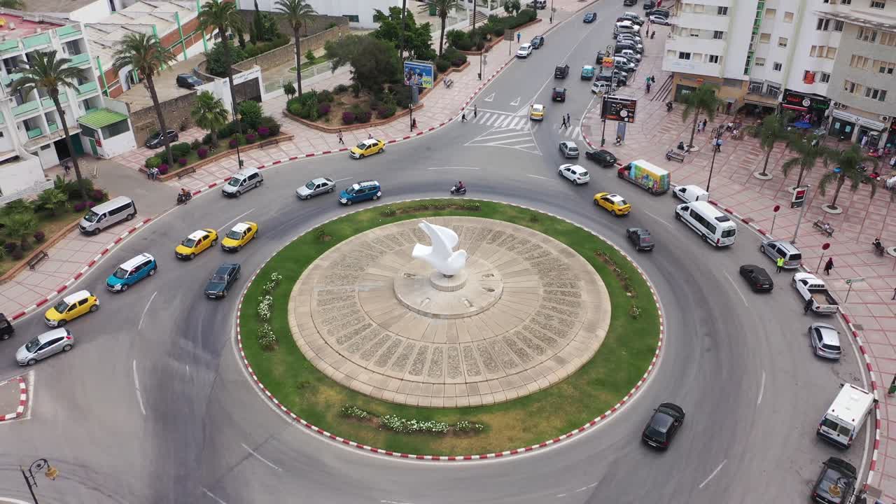 Una dinamica ripresa con drone che cattura il cuore di Tetouan, Marocco. Mostra il famoso monumento della Colomba Bianca, il traffico urbano vivace con taxi gialli e un'architettura mediterranea pulita e moderna.