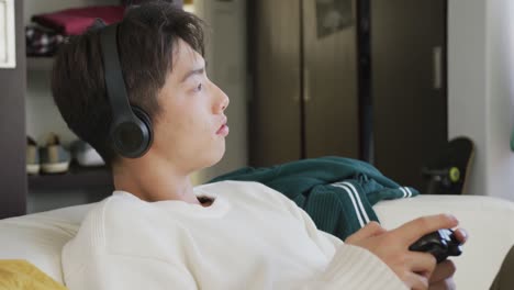 Asian-boy-wearing-headphones-playing-video-games-sitting-on-the-couch-at-home